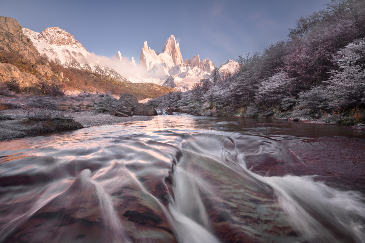 Water flows over red rock toward the camera, with frosted trees on the right and the snowy peaks of Fitz Roy behind.