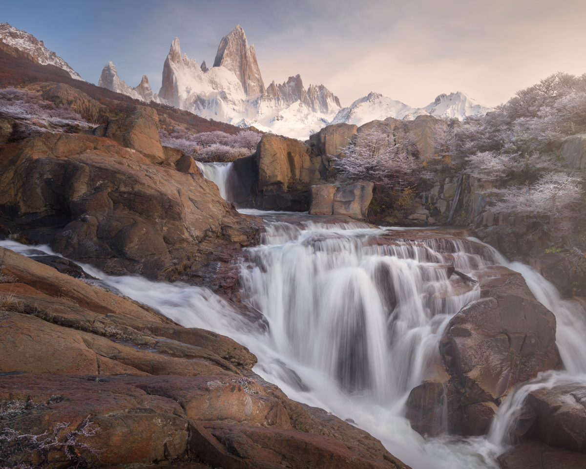 A wide waterfall flows over red rock between frosted shrubs, with the snowy peaks of Fitz Roy above in soft morning light.