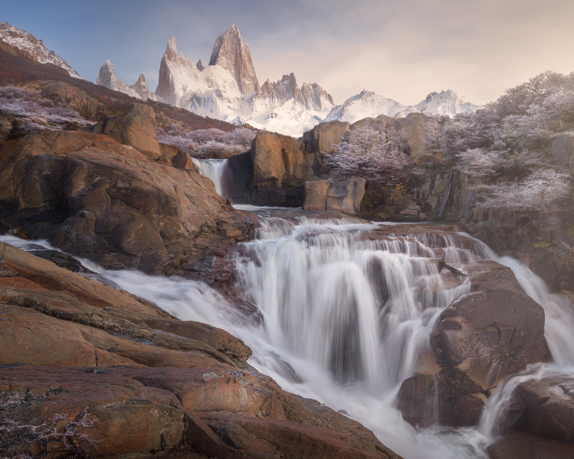 A wide waterfall flows over red rock between frosted shrubs, with the snowy peaks of Fitz Roy above in soft morning light.