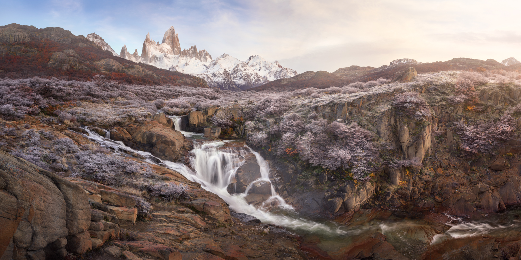 A wide view of small waterfalls in a rocky gorge, with frosted shrubs on the slopes and Fitz Roy on the left under a pale sky.