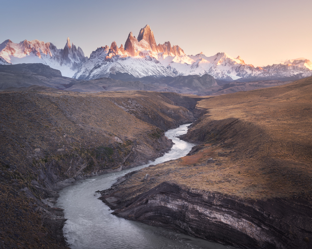The Rio de las Vueltas bends through a canyon with layered rock walls, below the snowy peaks of Fitz Roy lit by the first sun.