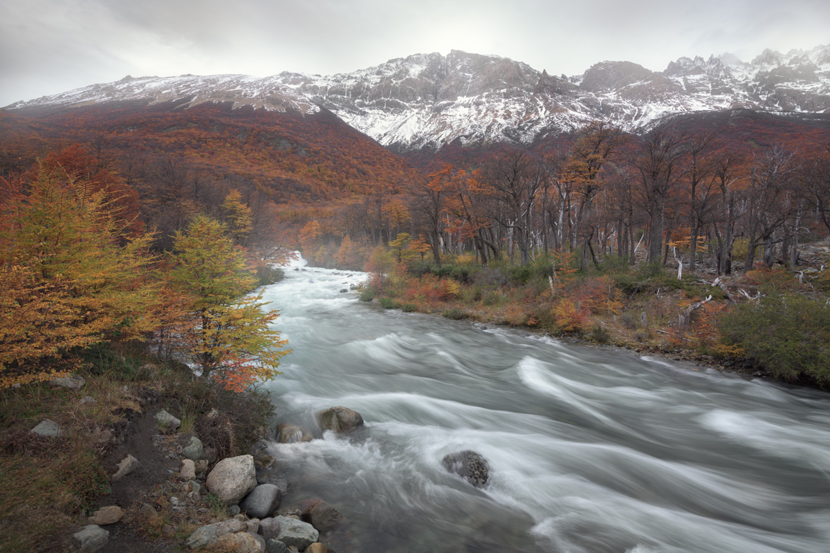A fast river runs between orange and yellow autumn trees, with large stones on the left bank and a snowy ridge under a grey sky.