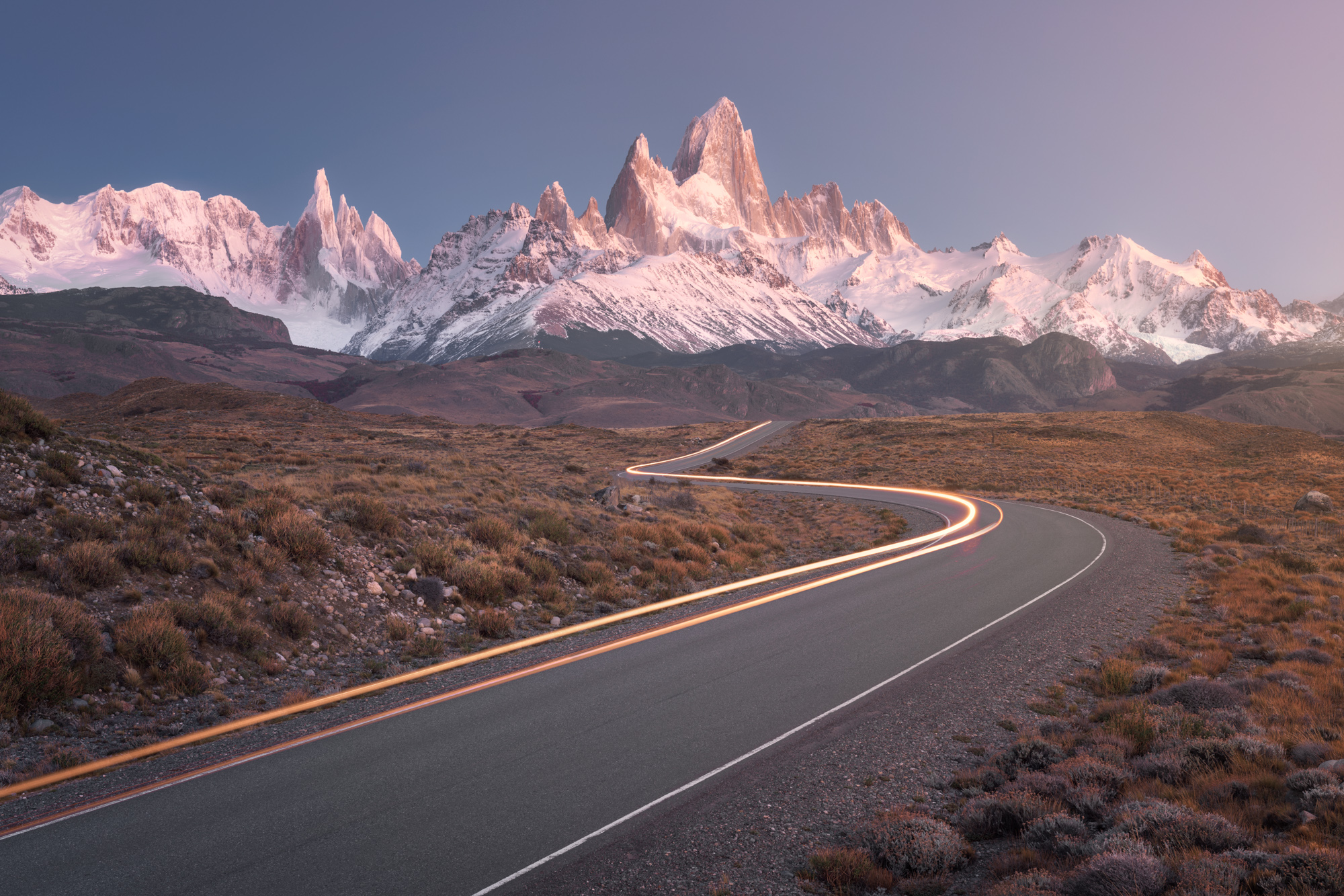 A curving road with an orange light trail leads toward the snowy peaks of Fitz Roy and Cerro Torre, lit pink at dawn.